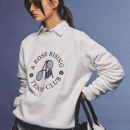 Arosris club white tennis club heritage sweatshirt with vintage crest graphic layered over a crisp collared shirt, elegantly styled on a model for an effortless 90s tenniscore and old money sporty rich aesthetic.
